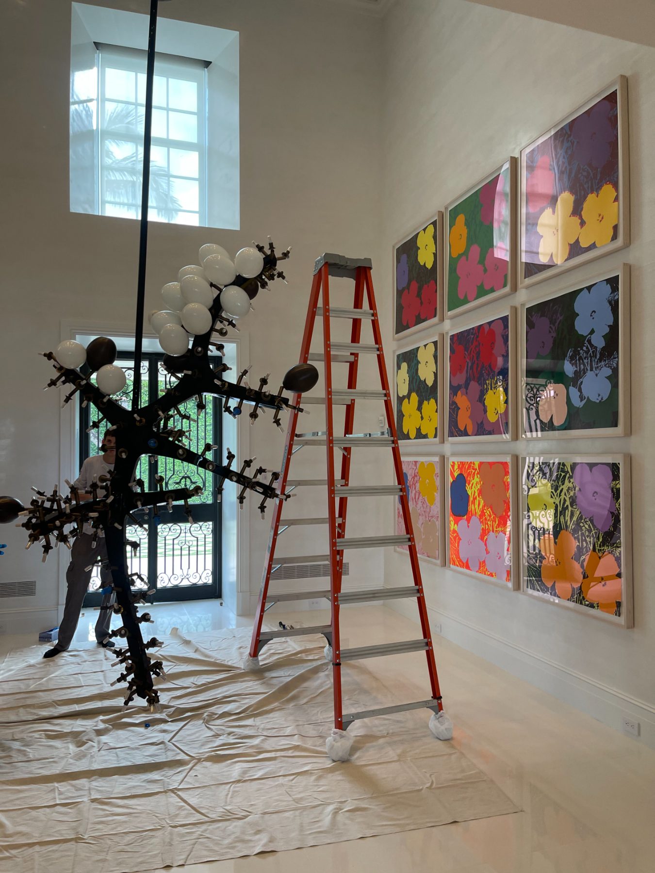 Installation in progress in a designer residence: framed prints hung in a grid and a sculptural light fixture on protective drop cloths